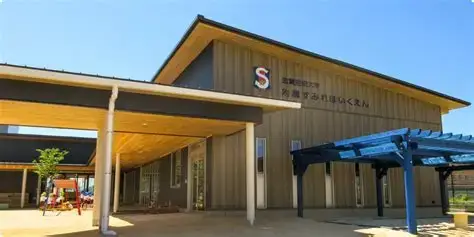 Exterior of a single-story public building with a tan facade, a sloped roof, and a covered entrance; Japanese signage near the top.