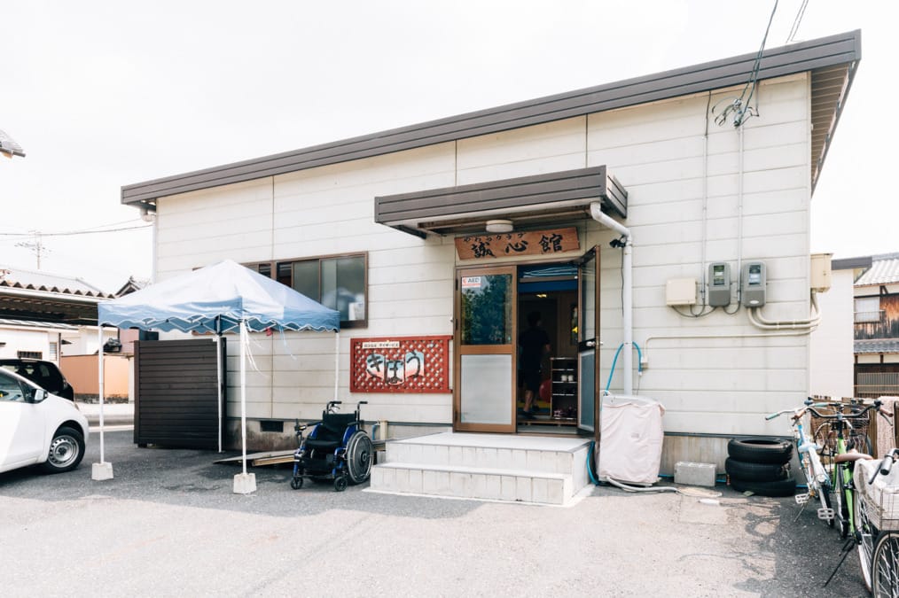 Exterior view of a small beige building with a wooden doorway and Japanese sign above the entrance; a wheelchair sits near the steps and a blue canopy tent is set up to the left outside. bicycles and stacked tires are visible on the right side of the image.