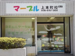 Front of a Japanese tutoring school storefront with a colorful sign above the entrance, glass doors with decorative white patterns, and a trimmed hedge in front.