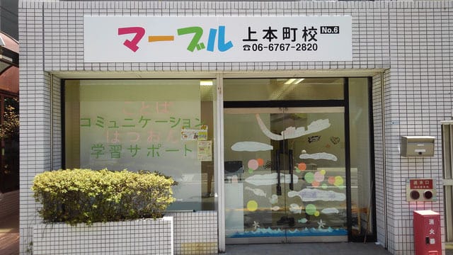 Front of a Japanese tutoring school storefront with a colorful sign above the entrance, glass doors with decorative white patterns, and a trimmed hedge in front.