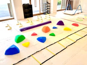 Bright therapy room with colorful soft play shapes, balance steps, and agility ladders for kids' activities on a sunny white floor.