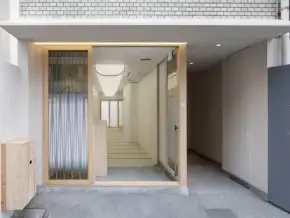 Modern hotel entrance with a wooden-framed glass doorway opening to a bright white interior hallway and stairs; exterior gray wall with the hotel name on the right.