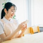 Young woman in a peach apron sits at a light wood table, smiling while looking at her phone.