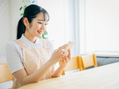Young woman in a peach apron sits at a light wood table, smiling while looking at her phone.