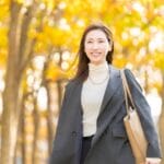 Smiling woman walking in a sunlit autumn park with yellow leaves, wearing a gray blazer, turtleneck, and carrying a beige tote bag.