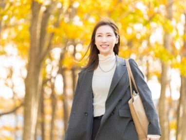Smiling woman walking in a sunlit autumn park with yellow leaves, wearing a gray blazer, turtleneck, and carrying a beige tote bag.