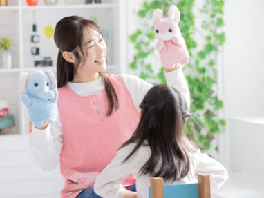Smiling woman in a pink apron uses blue elephant and pink bunny hand puppets to play with a child in a bright room.