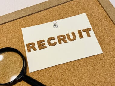 Recruitment sign pinned to a corkboard with a magnifying glass nearby, suggesting job openings.