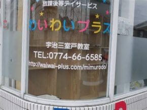 Storefront window advertising Wai Wai Plus after-school day service, with colorful Japanese text and contact info (TEL 0774-66-6585) and a website URL.