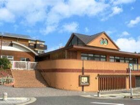 Tan and brown brick building with a curved roof and a logo on the gable, set against a blue sky; stairs lead to the entrance.