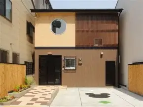 Front view of a modern two-story house with brown siding, a circular window on the upper left, and a small garden bed beside a checkered walkway.