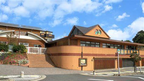 Tan and brown brick building with a curved roof and a logo on the gable, set against a blue sky; stairs lead to the entrance.