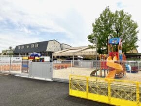 Outdoor fenced playground with a bright yellow spiral slide, colorful play structures, and a shaded seating area near a gray building under a partly cloudy sky.