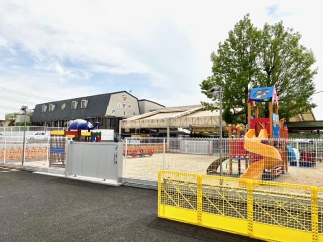 Outdoor fenced playground with a bright yellow spiral slide, colorful play structures, and a shaded seating area near a gray building under a partly cloudy sky.