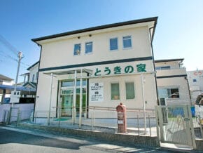 Two-story white building with green Japanese signage (とおきの家), glass entry, and a red mailbox outside on a sunny day.
