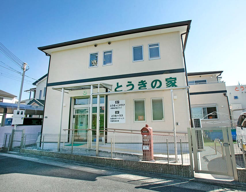 Two-story white building with green Japanese signage (とおきの家), glass entry, and a red mailbox outside on a sunny day.