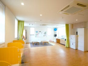 Bright classroom/playroom with low benches, rug, whiteboard, and a kitchenette at the back.