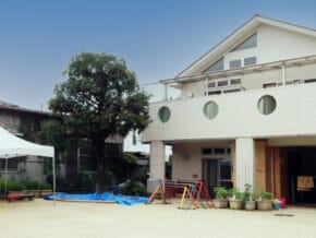 A light-colored two-story building with circular windows, a small playground with a set of colorful bars, and a white canopy tent in a yard.