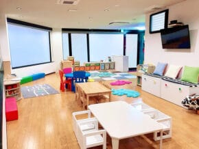 Bright, spacious children's playroom with kid-sized white tables and chairs, colorful cushions, storage cubbies, and a wall-mounted TV.
