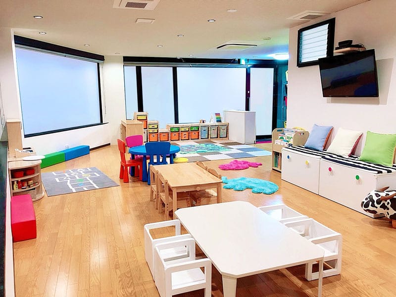 Bright, spacious children's playroom with kid-sized white tables and chairs, colorful cushions, storage cubbies, and a wall-mounted TV.