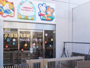 Front entrance of a Japanese daycare center with glass doors, colorful decals, and cartoon animal murals above the doorway on a tiled exterior wall.