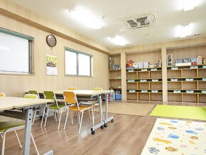 Bright classroom with empty tables and colorful chairs, and open storage shelves along the back wall.