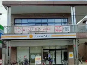 Two-story storefront building with 'chocoZAP' sign on the ground level and bicycles parked at the entrance, a balcony above with banners in Japanese.