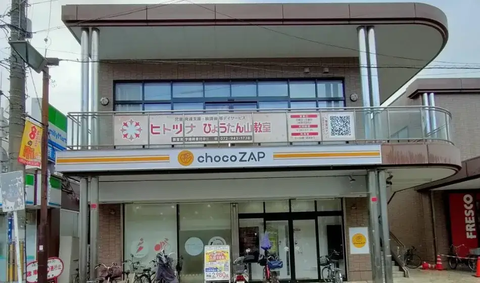 Two-story storefront building with 'chocoZAP' sign on the ground level and bicycles parked at the entrance, a balcony above with banners in Japanese.