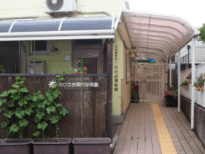 Entrance to a building with a curved translucent canopy, brick walkway, and potted plants along a wooden fence; a yellow tactile paving strip runs down the center for accessibility.