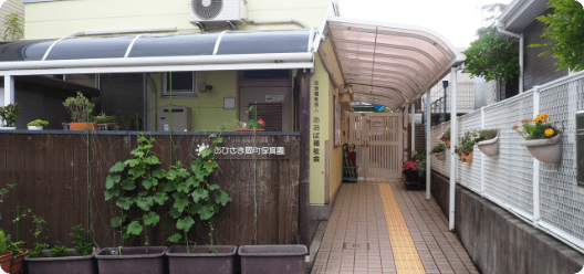 202407301725205518 | 保育士求人|大阪・兵庫・関西の保育士・幼稚園教諭の就職転職支援|保育のせかい Entrance to a building with a curved translucent canopy, brick walkway, and potted plants along a wooden fence; a yellow tactile paving strip runs down the center for accessibility.