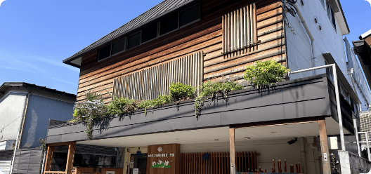 Two-story wooden storefront with a raised planter box and green shrubs, under a clear blue sky.