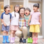 A smiling female teacher kneels with four children outside, all wearing colorful rain boots and casual clothes, posing for a group photo.