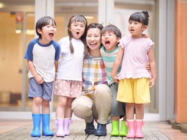 A smiling female teacher kneels with four children outside, all wearing colorful rain boots and casual clothes, posing for a group photo.