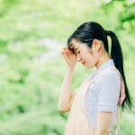 Young woman with a ponytail standing in a green outdoor setting, touching her forehead as if tired or sad.