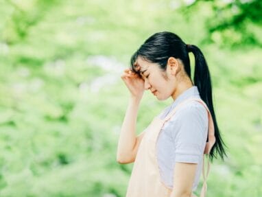 Young woman with a ponytail standing in a green outdoor setting, touching her forehead as if tired or sad.