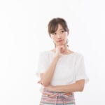 Young woman in a white shirt stands with arms crossed, resting her chin on her hand in a thoughtful pose.