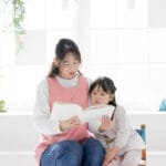 Two young girls sit together reading a book in a bright, plant-filled room.