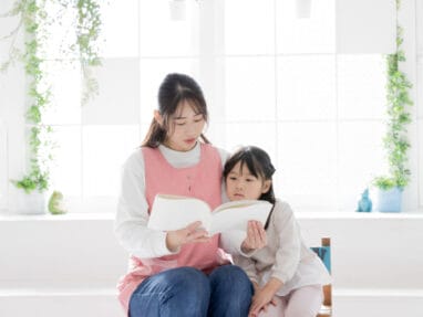 Two young girls sit together reading a book in a bright, plant-filled room.