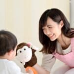 Woman in a pink apron smiles at a small child, presenting a brown plush monkey toy during playtime.