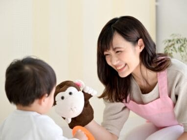 4211867_s | 保育士求人|大阪・兵庫・関西の保育士・幼稚園教諭の就職転職支援|保育のせかい Woman in a pink apron smiles at a small child, presenting a brown plush monkey toy during playtime.