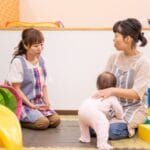 Two caregivers in a playroom talk while a baby stands beside the seated caregiver.