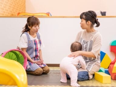 Two caregivers in a playroom talk while a baby stands beside the seated caregiver.