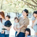 Group of smiling moms with babies and young children standing together in a sunlit park.
