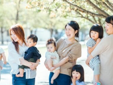 Group of smiling moms with babies and young children standing together in a sunlit park.