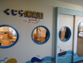 Wall of a Japanese daycare with blue circular windows; classrooms visible inside, sign reads くじら保育園.
