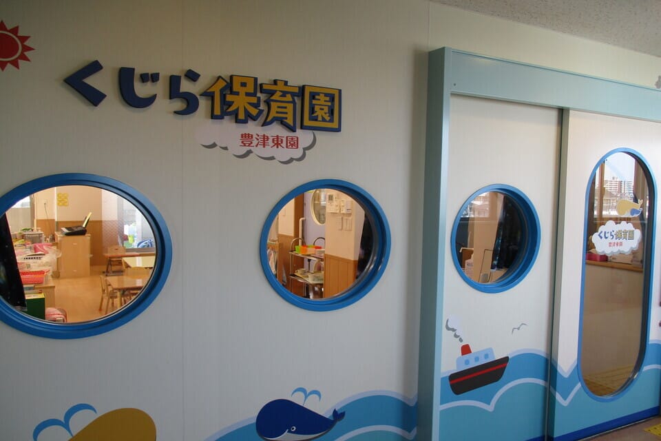 Wall of a Japanese daycare with blue circular windows; classrooms visible inside, sign reads くじら保育園.