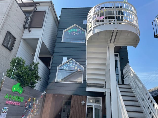 Two-story children's daycare building with a white external staircase, blue-gray siding, and windows decorated with colorful stickers.