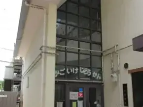 Entrance to a beige building with tall glass front and doors covered in colorful stickers; exposed pipes run along the exterior wall.