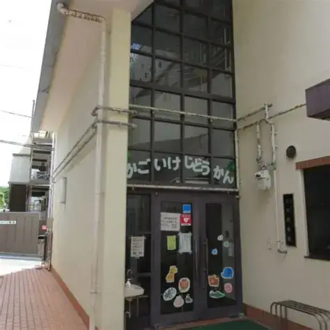 OIP 5 | 保育士求人|大阪・兵庫・関西の保育士・幼稚園教諭の就職転職支援|保育のせかい Entrance to a beige building with tall glass front and doors covered in colorful stickers; exposed pipes run along the exterior wall.
