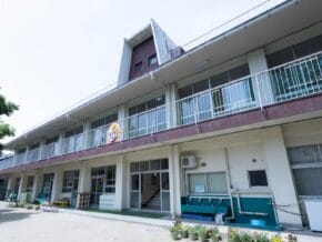 Two-story building with balconies and a small outdoor play area to the left; potted plants line the front.